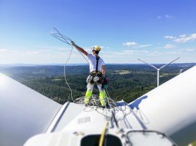 Rear view on Professional rope access technician standing on roof (hub) of wind turbine and pulling rope up. Sun is behind wind turbine.